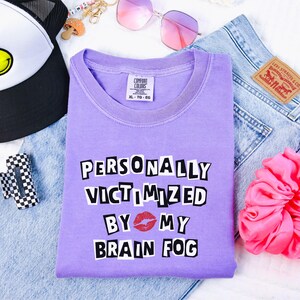 Puede incluir: Una camiseta lila Comfort Colors con el texto "PERSONALLY VICTIMIZED BY MY BRAIN FOG" en negrita, en blanco y negro, con un gráfico de beso de lápiz labial rojo. La camiseta está colocada sobre unos vaqueros, con un accesorio a cuadros en blanco y negro, una gorra de camionero y gafas de sol rosas.