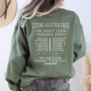 May include: Olive green sweatshirt with the text "LIVING GLUTEN-FREE THE DAILY FOOD STRUGGLE TOUR" in white. The back of the sweatshirt lists common challenges of a gluten-free diet. The text includes phrases like "Bloating & Pain" and "Tummy Ache Survivor".