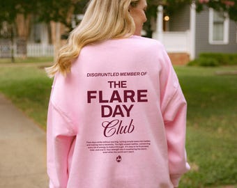 May include: Pink sweatshirt with the text "DISGRUNTLED MEMBER OF THE FLARE DAY Club" in maroon lettering. The text is centered on the back of the sweatshirt. The text below the club name reads "Flare days strike without warning, turning simple tasks into battles and making rest a necessity. You fight unseen battles, remembering every bit of energy to make it through. You're weathering the storm, tired, and over it. Your strength lies in weathering the storm, even when the world can't see it."