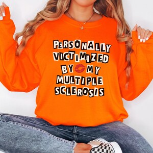 Könnte beinhalten: Leuchtend orangefarbenes Sweatshirt mit dem Text "PERSONALLY VICTIMIZED BY MY MULTIPLE SCLEROSIS" in einem schwarz-weißen Blockbuchstaben-Design. Das Wort "BY" hat eine rote Lippen-Grafik. Das Sweatshirt hat lange Ärmel.