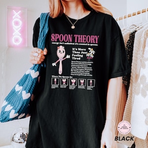 May include: A black t-shirt featuring a pink cartoon spoon character and the text Spoon Theory, energy isn't unlimited, it's counted in spoons. It includes a diagram explaining energy levels with small spoon icons and descriptive text.