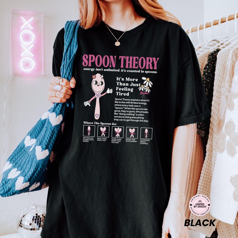 May include: A black t-shirt featuring a pink cartoon spoon character and the text Spoon Theory, energy isn't unlimited, it's counted in spoons. It includes a diagram explaining energy levels with small spoon icons and descriptive text.