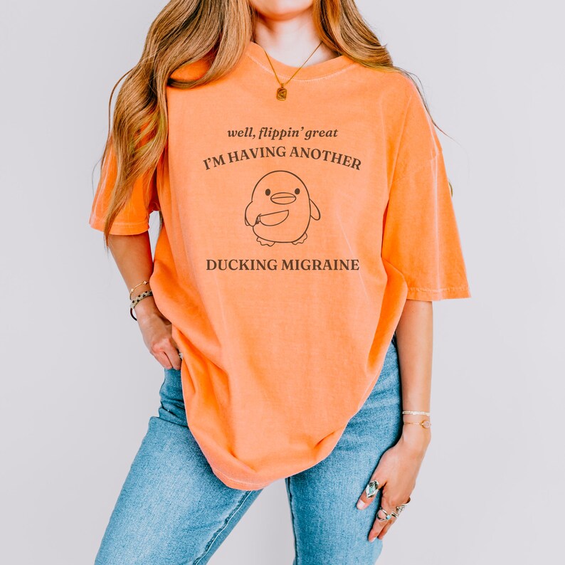 May include: A bright orange t-shirt with a graphic of a duck with the text "well, flippin' great I'M HAVING ANOTHER DUCKING MIGRAINE"
