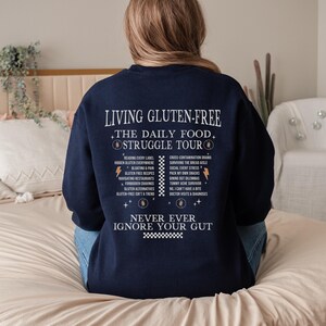 May include: Navy blue sweatshirt with white text that reads "LIVING GLUTEN-FREE THE DAILY FOOD STRUGGLE TOUR" and a list of related topics. The phrase "NEVER EVER IGNORE YOUR GUT" is also printed on the sweatshirt.