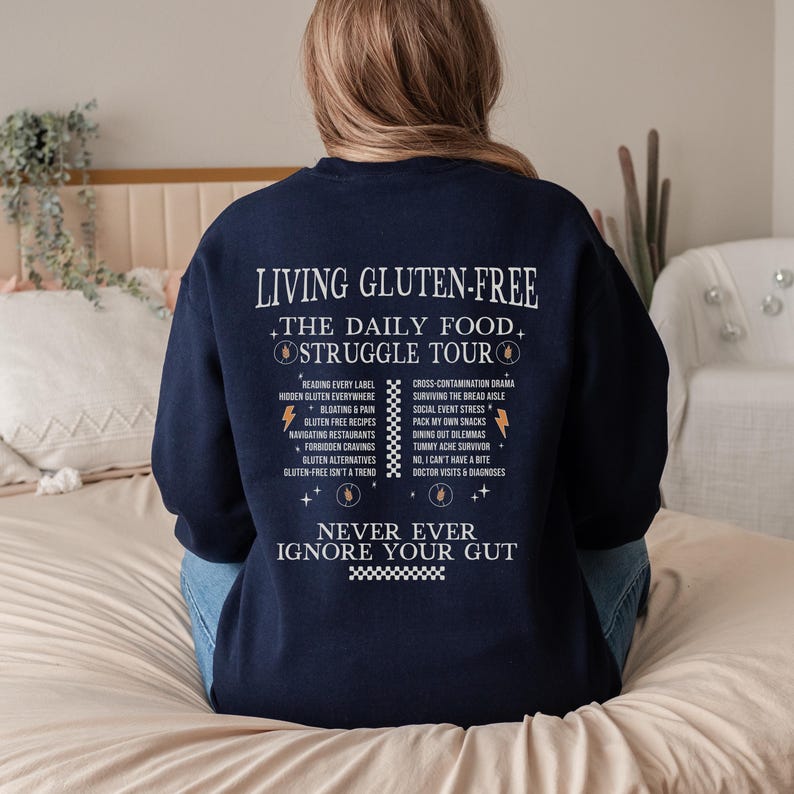 May include: Navy blue sweatshirt with white text that reads "LIVING GLUTEN-FREE THE DAILY FOOD STRUGGLE TOUR" and a list of related topics. The phrase "NEVER EVER IGNORE YOUR GUT" is also printed on the sweatshirt.