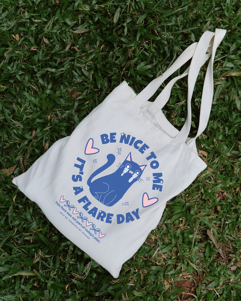 May include: White tote bag with blue and pink text that reads "Be Nice To Me It's A Flare Day". A blue cartoon cat with pink hearts and stars is crying. The text "This flare was not part of today's plan. Help me conserve my spoons" is printed below the cat.