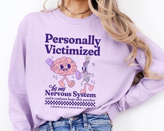 May include: Purple long-sleeve sweatshirt with a graphic of a brain and a skeleton boxing. The text reads "Personally Victimized by my Nervous System and it confuses hugs with punches. Fibromyalgia Awareness Club."