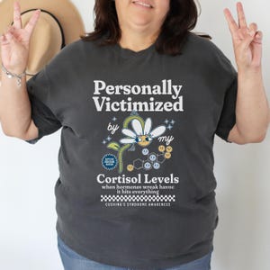 May include: A dark gray t-shirt with a white graphic design that says "Personally Victimized by my Cortisol Levels" with a cartoon daisy and a chemical structure. The text below the graphic says "when hormones wreak havoc it hits everything" and "Cushing's Syndrome Awareness".