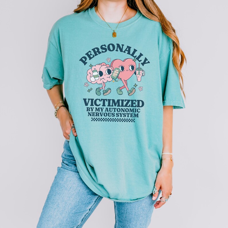 K&ouml;nnte beinhalten: Ein t&uuml;rkisfarbenes T-Shirt mit einer Grafik eines Gehirns und eines Herzens, die eine Tasse Kaffee halten. Der Text auf dem Shirt lautet "PERSONALLY VICTIMIZED BY MY AUTONOMIC NERVOUS SYSTEM".