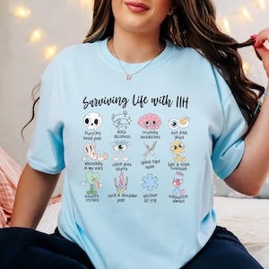 Może przedstawiać: Jasnoniebieski t-shirt z napisem Surviving Life with IIH oraz siatką dwunastu kolorowych ilustracji przedstawiających objawy, w tym czaszkę, mózg, oko, słońce i symbole medyczne wraz z opisowymi etykietami pod każdą z ikon.