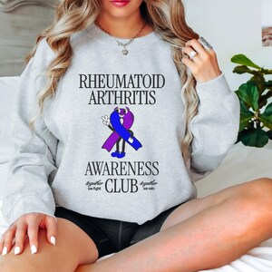 May include: A gray sweatshirt with the text "Rheumatoid Arthritis Awareness Club" and a purple and blue ribbon with a smiling face. The text "Together we fight, together we win" is below the ribbon.