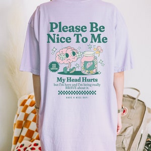 May include: Purple t-shirt with a graphic of a brain holding a jar with a heart and the text "Please Be Nice To Me", "Head Ache Survivor", "My Head Hurts but I'm here and I'm being really BRAVE about it. HAVE A NICE DAY!"
