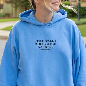 K&ouml;nnte beinhalten: Ein hellblauer Hoodie mit dem Aufdruck "Still Sweet Diabetes Warrior" auf der Vorderseite.