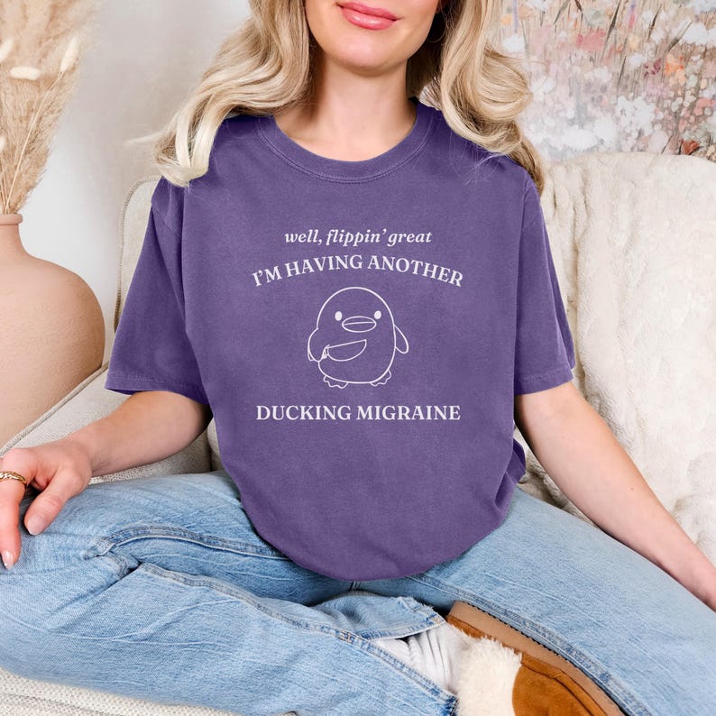 May include: Purple t-shirt with a white graphic of a penguin holding a knife and the text "well, flippin' great I'M HAVING ANOTHER DUCKING MIGRAINE"