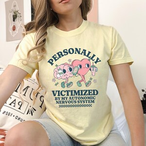 K&ouml;nnte beinhalten: Ein gelbes T-Shirt mit einer Grafik eines Gehirns und eines Herzens, die sich die H&auml;nde halten. Der Text auf dem Shirt lautet "PERSONALLY VICTIMIZED BY MY AUTONOMIC NERVOUS SYSTEM".