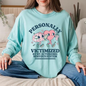 Puede incluir: Una camiseta de manga larga turquesa con un gráfico de un cerebro y un corazón sosteniendo bebidas. El texto dice "PERSONALLY VICTIMIZED BY MY AUTONOMIC NERVOUS SYSTEM".