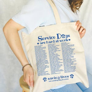 Könnte beinhalten: Eine beige Tragetasche aus Segeltuch mit blauem Text, der Service Dogs are hard at work lautet, gefolgt von einer detaillierten Liste von Assistenzhund-Aufgaben und dem Satz saving lives one working day at a time mit Pfotenabdruck-Symbolen.