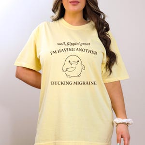 May include: A yellow t-shirt with the text "well, flippin' great I'M HAVING ANOTHER DUCKING MIGRAINE" and a cartoon duck with a knife.