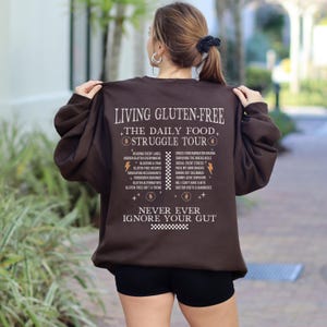 May include: A brown sweatshirt with the text "LIVING GLUTEN-FREE THE DAILY FOOD STRUGGLE TOUR" and a list of related topics. The sweatshirt also has the text "NEVER EVER IGNORE YOUR GUT".
