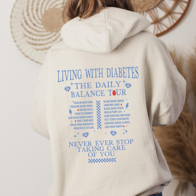 K&ouml;nnte beinhalten: Ein cremefarbener Hoodie mit einem blauen und roten Grafikdruck, der "Leben mit Diabetes + Die t&auml;gliche Balance-Tour" sagt und h&auml;ufige Herausforderungen des Lebens mit Diabetes auflistet. Die Grafik enth&auml;lt auch den Text "H&ouml;r niemals auf, auf dich aufzupassen".