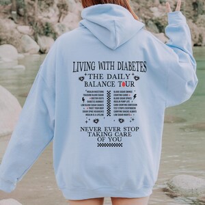 K&ouml;nnte beinhalten: Hellblauer Hoodie mit einem schwarzen Grafikdruck, der "Leben mit Diabetes + Die t&auml;gliche Balance-Tour" lautet und verschiedene Aspekte der Diabetes-Behandlung auflistet. Die Grafik enth&auml;lt auch den Text "H&ouml;r niemals auf, auf dich aufzupassen".