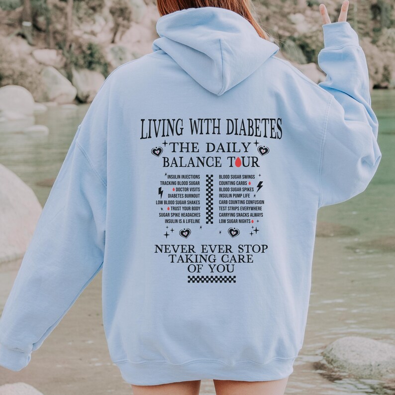 K&ouml;nnte beinhalten: Hellblauer Hoodie mit einem schwarzen Grafikdruck, der "Leben mit Diabetes + Die t&auml;gliche Balance-Tour" lautet und verschiedene Aspekte der Diabetes-Behandlung auflistet. Die Grafik enth&auml;lt auch den Text "H&ouml;r niemals auf, auf dich aufzupassen".