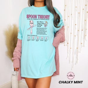 May include: A light blue t-shirt featuring a pink spoon character graphic and the text Spoon Theory, energy isn't unlimited, it's counted in spoons. It includes a diagram explaining the theory with small spoon icons and descriptive text.