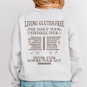 May include: A light gray sweatshirt with the text "LIVING GLUTEN-FREE" and "THE DAILY FOOD STRUGGLE TOUR" in brown. The back of the sweatshirt lists common challenges of a gluten-free diet. The text "NEVER EVER IGNORE YOUR GUT" is also printed.