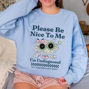 Puede incluir: Sudadera azul claro con el texto "Please Be Nice To Me" y "I'm Undiagnosed". El gráfico presenta un diseño caprichoso de un personaje parecido a un fideo con gafas de sol y un lazo. También incluye la frase "BUT SOMETHING AIN'T RIGHT".