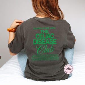 Op de afbeelding: Donkergrijs T-shirt met de groene tekst "GLUTEN IS NOT SAFE. THE CELIAC DISEASE CLUB". Het shirt heeft ook een beschrijving van Coeliakie in het groen. Het shirt heeft een comfortabele pasvorm.