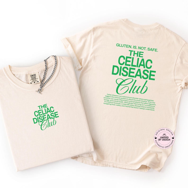 Op de afbeelding: Twee cr&egrave;mekleurige t-shirts met groene tekst "THE CELIAC DISEASE Club". E&eacute;n shirt is opgevouwen, de andere is uitgevouwen en toont de achterkant met de tekst "GLUTEN. IS. NOT. SAFE." en een beschrijving van coeliakie.