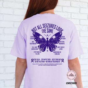 Op de afbeelding: Een lichtpaars t-shirt met een donkerpaarse afbeelding op de achterkant. Het ontwerp bevat een vlinder en de tekst "NOT ALL SEIZURES LOOK THE SAME" samen met een lijst van symptomen van aanvallen en veiligheidsinstructies voor hulpverlening.