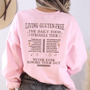 May include: A light pink sweatshirt with the text "LIVING GLUTEN-FREE THE DAILY FOOD STRUGGLE TOUR" in a dark brown color. The back of the sweatshirt lists common challenges of a gluten-free diet. The phrase "NEVER EVER IGNORE YOUR GUT" is also printed on the sweatshirt.