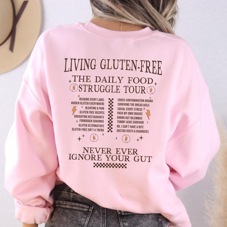 May include: A light pink sweatshirt with the text "LIVING GLUTEN-FREE THE DAILY FOOD STRUGGLE TOUR" in a dark brown color. The back of the sweatshirt lists common challenges of a gluten-free diet. The phrase "NEVER EVER IGNORE YOUR GUT" is also printed on the sweatshirt.