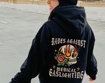 May include: Black hooded sweatshirt with a skull and rose graphic and the text "Babes Against Medical Gaslighting".
