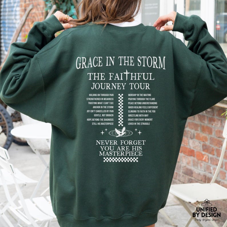 以下が含まれることがあります： 白い文字で「Grace in the Storm, The Faithful Journey Tour」とフレーズのリストが書かれた濃い緑色のスウェットシャツ。スウェットシャツの下部には「Never Forget You Are His Masterpiece」という文字があります。
