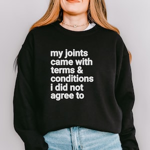 Könnte beinhalten: Schwarzes Sweatshirt mit dem weißen Text "my joints came with terms & conditions i did not agree to." Das Sweatshirt wird von einer Person mit langen blonden Haaren und einer Jeans getragen.