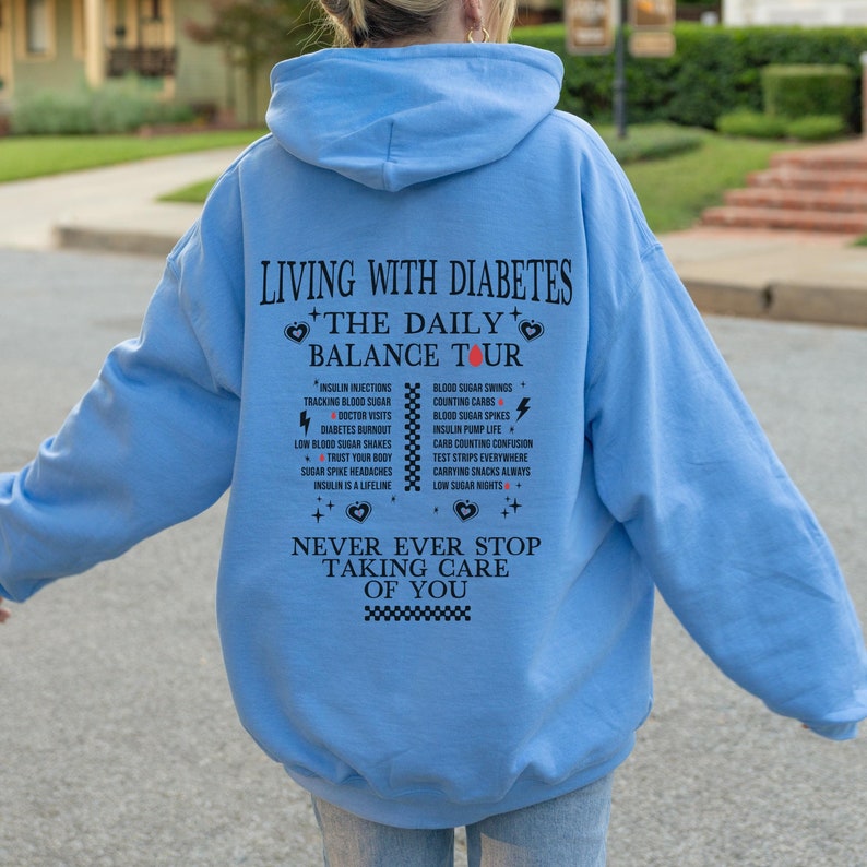 K&ouml;nnte beinhalten: Hellblauer Kapuzenpullover mit dem Text "Leben mit Diabetes + Die t&auml;gliche Balance-Tour" und einer Liste von t&auml;glichen Aufgaben im Zusammenhang mit der Diabetes-Behandlung. Der Text "H&ouml;r niemals auf, auf dich aufzupassen" ist unter der Liste gedruckt.