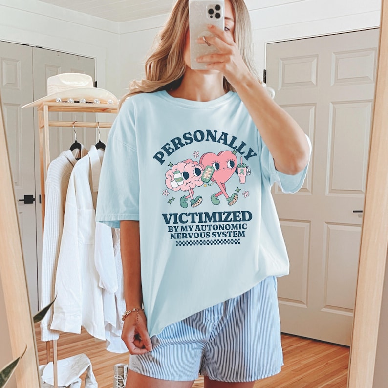 K&ouml;nnte beinhalten: Ein hellblaues T-Shirt mit einer Grafik eines Gehirns und eines Herzens, die Getr&auml;nke halten. Der Text auf dem Shirt lautet "PERSONALLY VICTIMIZED BY MY AUTONOMIC NERVOUS SYSTEM".