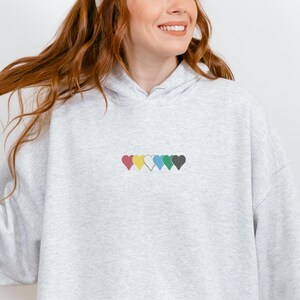 Peut inclure: Un sweat à capuche gris clair orné d'une rangée de six cœurs colorés brodés sur la poitrine. Les cœurs sont roses, jaunes, blancs, bleus, verts et gris. Le sweat à capuche a une capuche à cordon et des manches longues.