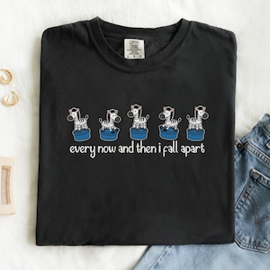 Könnte beinhalten: Schwarzes T-Shirt mit einem verspielten Design von fünf Zebras auf blauen Podesten. Die Zebras sind in einer Reihe angeordnet, darunter der Text "every now and then I fall apart". Das Shirt ist von der Marke Comfort Colors.