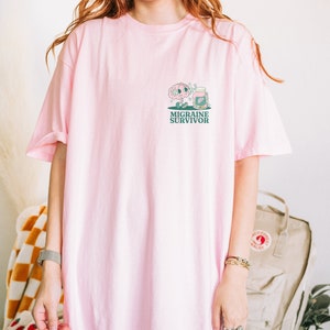 May include: Pink oversized t-shirt with a cartoon brain and pill bottle graphic and the text "Migraine Survivor".