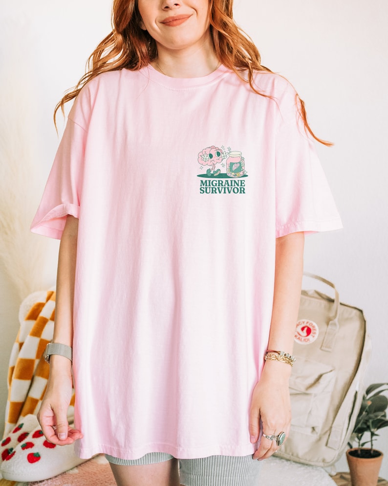 May include: Pink oversized t-shirt with a cartoon brain and pill bottle graphic and the text "Migraine Survivor".