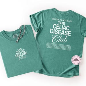 Op de afbeelding: Twee groene t-shirts met witte tekst "THE CELIAC DISEASE Club". E&eacute;n shirt heeft een zilveren ketting. De achterkant van &eacute;&eacute;n shirt leest "GLUTEN. IS. NOT. SAFE." en heeft een beschrijving van Coeliakie.