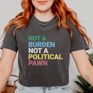 May include: A dark gray cropped t-shirt with the text "NOT A BURDEN NOT A POLITICAL PAWN" in colorful letters.
