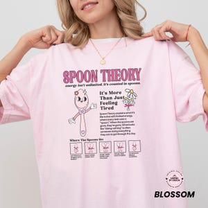 May include: Pink t-shirt with the text "SPOON THEORY" and an illustration of a spoon character. The shirt also includes text explaining the Spoon Theory concept and the phrase "It's More Than Just Feeling Tired."