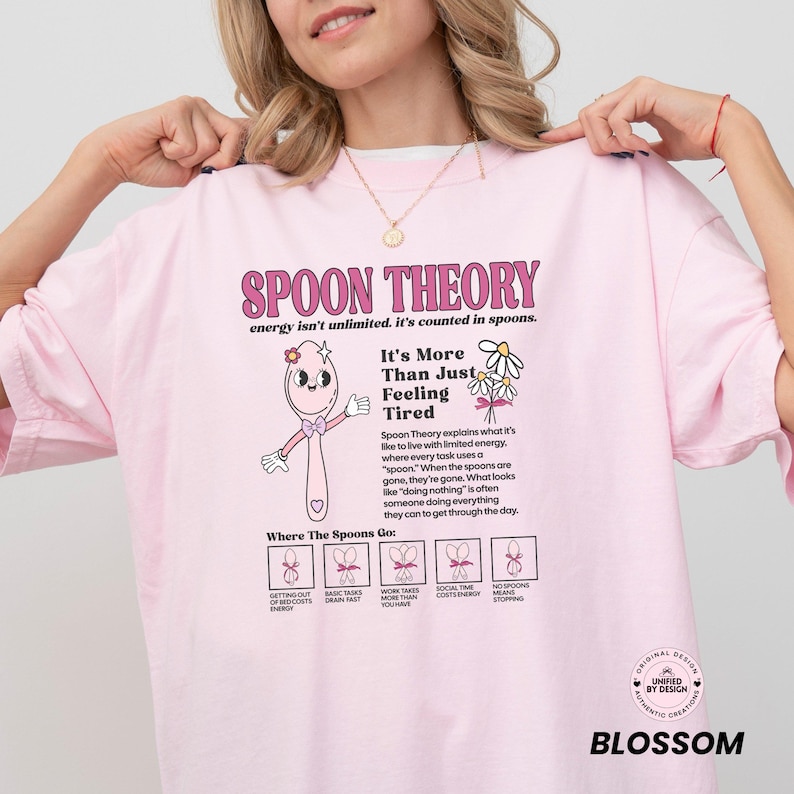 May include: Pink t-shirt with the text "SPOON THEORY" and an illustration of a spoon character. The shirt also includes text explaining the Spoon Theory concept and the phrase "It's More Than Just Feeling Tired."
