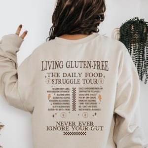 Puede incluir: Sudadera beige con cuello redondo con el texto "LIVING GLUTEN-FREE" y una lista de problemas sin gluten en marrón oscuro. La frase "NEVER EVER IGNORE YOUR GUT" está impresa en la parte inferior. Una prenda cómoda e informal.