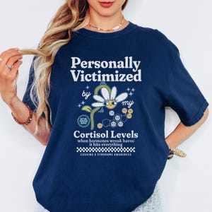 May include: Navy blue t-shirt with a daisy graphic and the text "Personally Victimized by my Cortisol Levels when hormones wreak havoc it hits everything Cushing's Syndrome Awareness".
