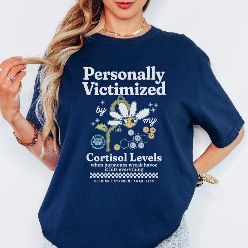 May include: Navy blue t-shirt with a daisy graphic and the text "Personally Victimized by my Cortisol Levels when hormones wreak havoc it hits everything Cushing's Syndrome Awareness".